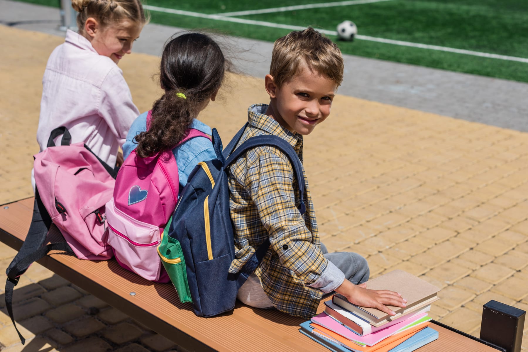 Schoolkids wear backpacks and sit on bench.