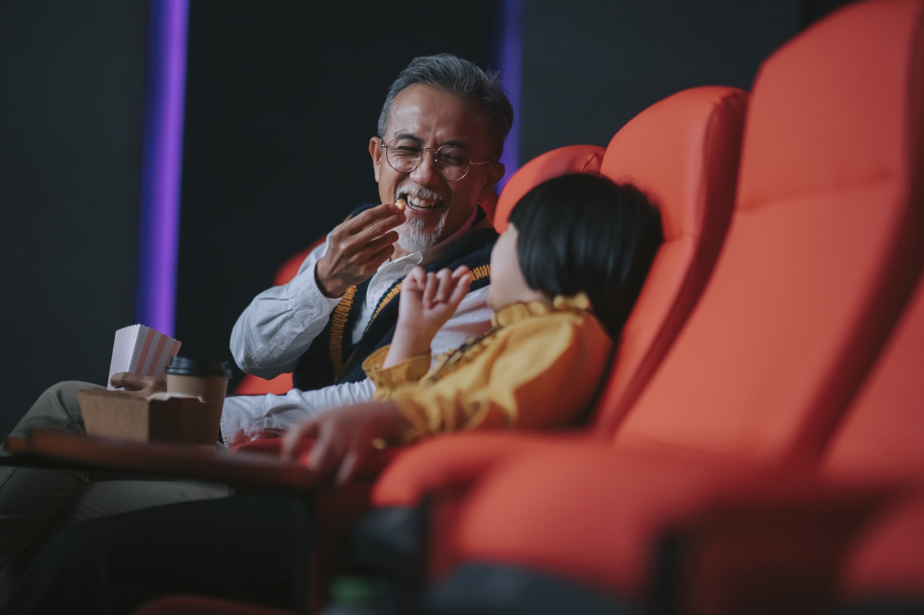 A man and his granddaughter enjoy snacks in a movie theater.