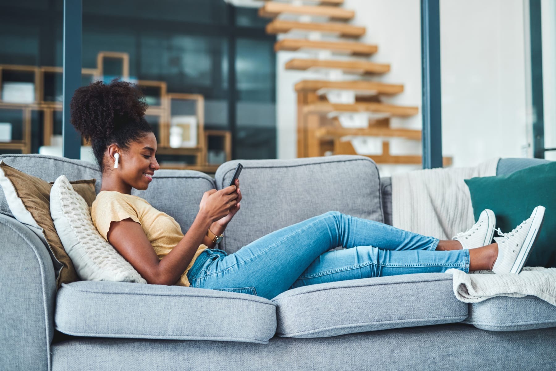 Young woman uses smartphone and wireless earbuds on sofa.