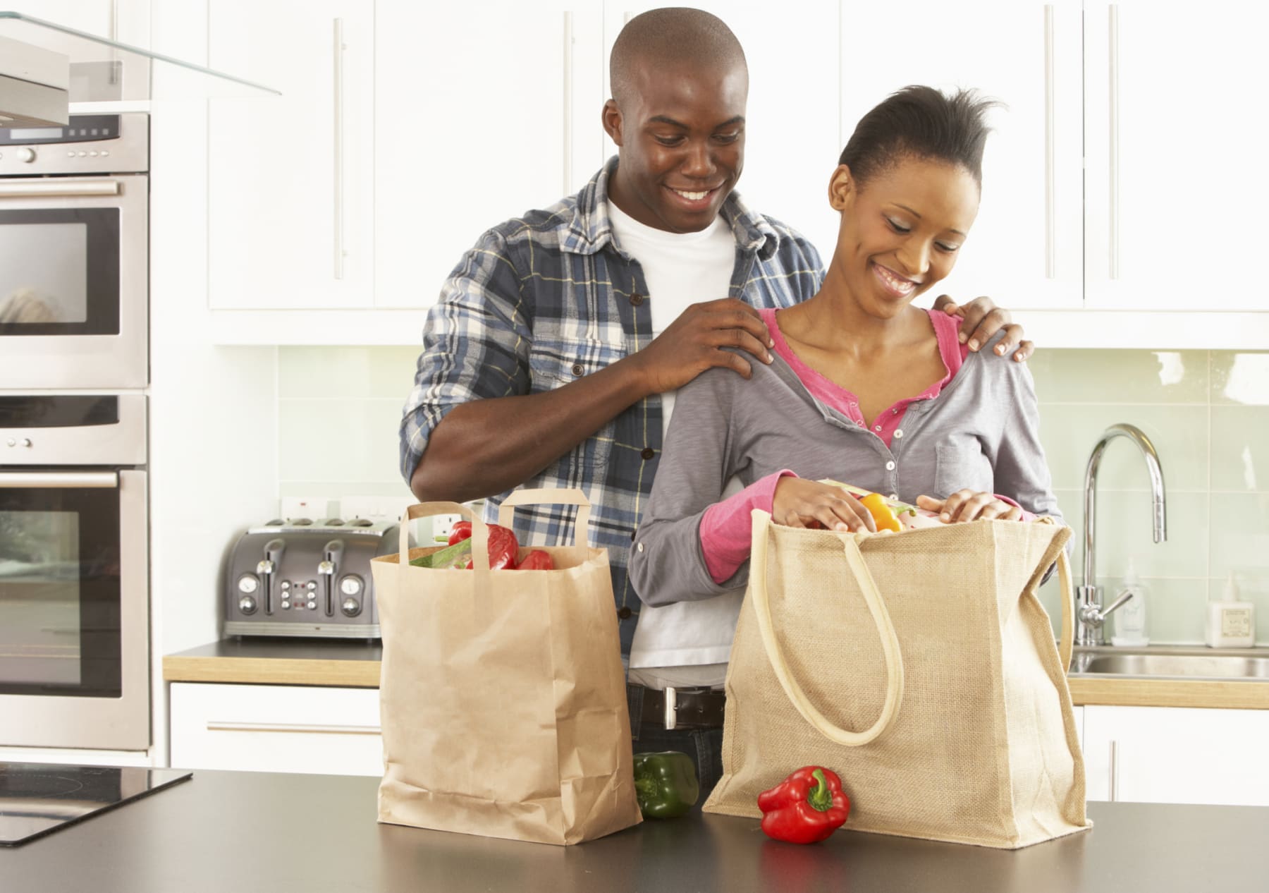 Couple stands in kitchen with bags full of food.